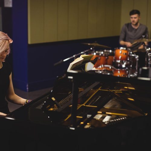 Female and male students playing piano and drum set in a studio